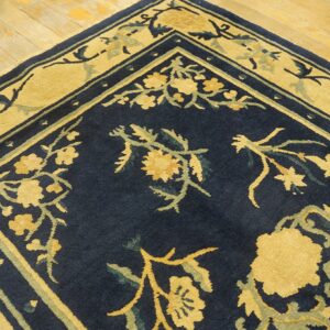 A corner of a navy blue rug with gold and sage floral patterns lies on light hardwood plank flooring.