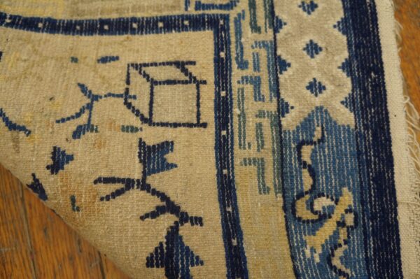 Low-pile tan rug corner displaying indigo and blue geometric motifs and a fringe edge on wood flooring.