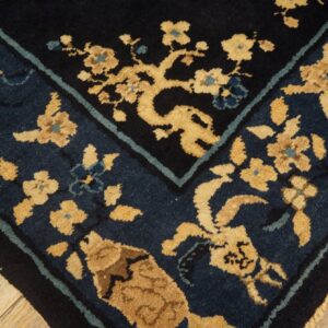 Dark blue and tan floral rug corner detail with a black border, visible over light wood flooring.