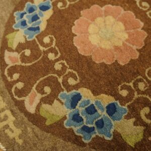 Brown circular rug with large pink and blue flowers, scrolling vines, and a geometric border on wood planks.