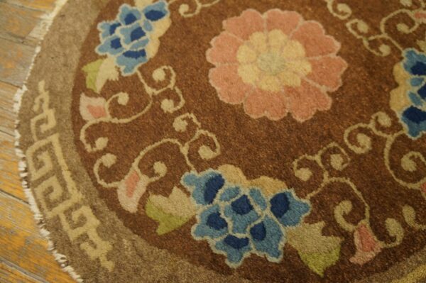 Brown circular rug with large pink and blue flowers, scrolling vines, and a geometric border on wood planks.