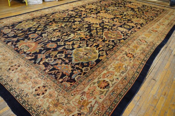 A large traditional rug with a navy center, colorful repeating motifs, and a wide beige border on wood floors.