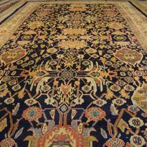 Navy blue rug with intricate gold and rust floral patterns and a wide beige border on wood floors.