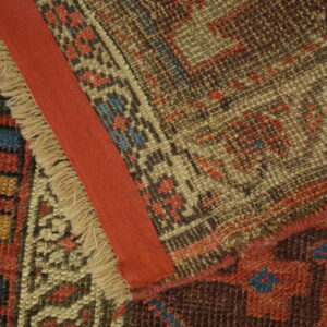 Geometric brown, beige, and blue rug folded to show knotted pile, frayed fringe, and a red fabric edge binding.