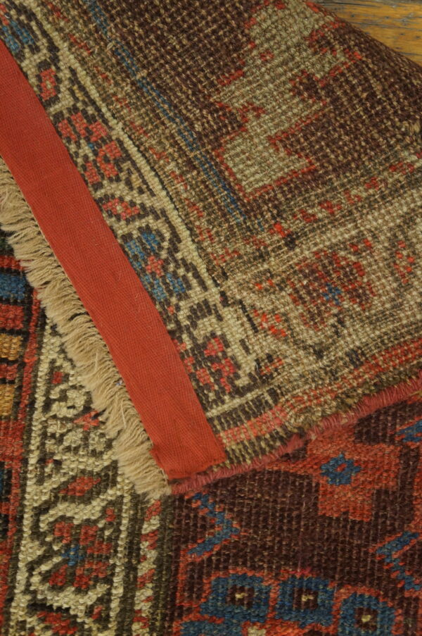 Geometric brown, beige, and blue rug folded to show knotted pile, frayed fringe, and a red fabric edge binding.