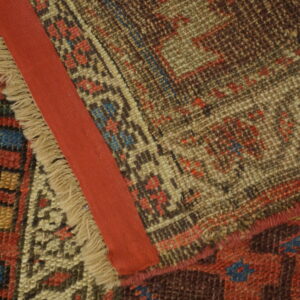Close-up of a geometrically patterned rug folded over, featuring fringe, a red fabric strip, and wooden flooring.