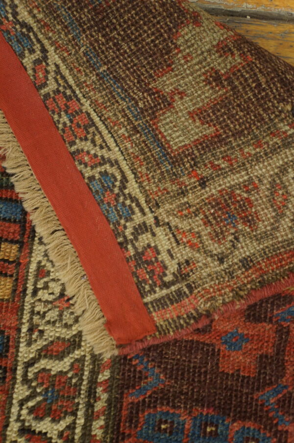 Close-up of a geometrically patterned rug folded over, featuring fringe, a red fabric strip, and wooden flooring.