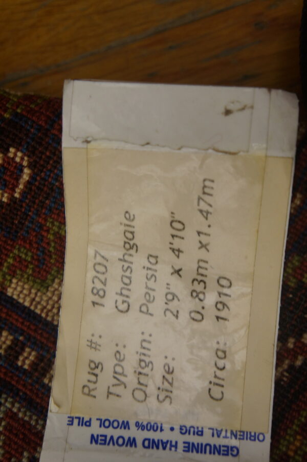 Low-pile geometric rug in red, blue, and cream with an attached label stating 100% wool pile.