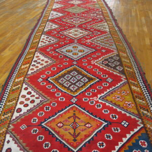 Vibrant red flatweave runner with complex multicolored geometric diamond patterns on light wood flooring.