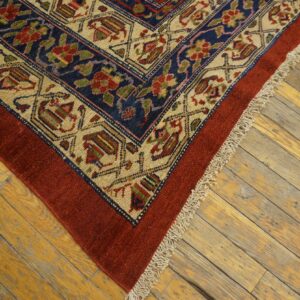 Red and navy patterned rug corner with fringe rests on rustic yellow wood planks.