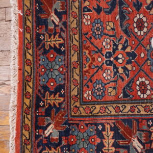 Close-up of a tightly woven rug with rust, blue, and gold patterns, featuring white fringe on hardwood floor.