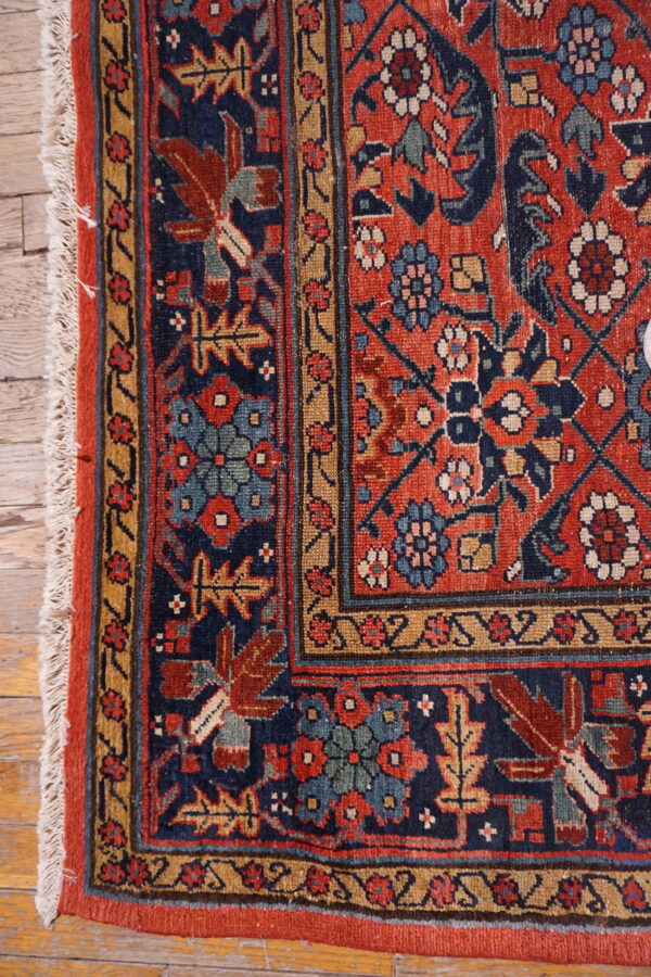 Close-up of a tightly woven rug with rust, blue, and gold patterns, featuring white fringe on hardwood floor.
