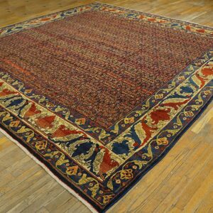 Square rug featuring a dense red field and wide navy border with stylized red and gold figures on wood flooring.