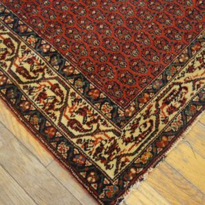 Red rug with a dense traditional pattern and cream border rests on light, worn hardwood planks.