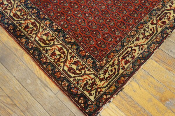 Red rug with a dense traditional pattern and cream border rests on light, worn hardwood planks.