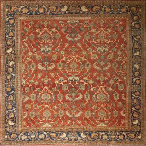 A detailed traditional rug featuring a rust red field with intricate floral patterns and a wide navy blue border.