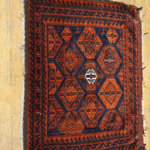 Square geometric rug in deep red-orange and navy blue on distressed wooden floorboards.