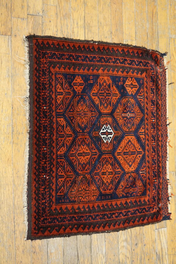 Square geometric rug in deep red-orange and navy blue on distressed wooden floorboards.