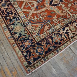 Rust rug with navy and beige border featuring complex geometric patterns rests on distressed gray wood planks.
