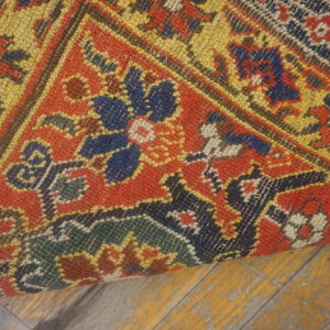 A curled rug section with red, yellow, blue, and green geometric motifs rests on worn yellow wooden flooring.