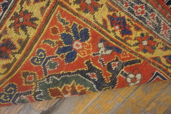 A curled rug section with red, yellow, blue, and green geometric motifs rests on worn yellow wooden flooring.