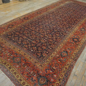 Traditional dark red, blue, and gold patterned rug displayed on rustic light wood flooring in a warehouse setting.