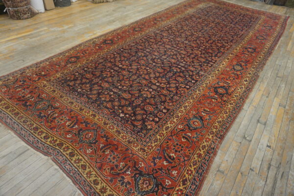 Traditional dark red, blue, and gold patterned rug displayed on rustic light wood flooring in a warehouse setting.