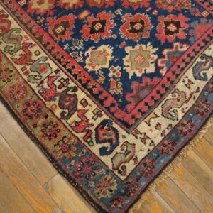 Low-pile tribal rug corner featuring blue, red, tan, and green patterns rests on wooden floorboards.