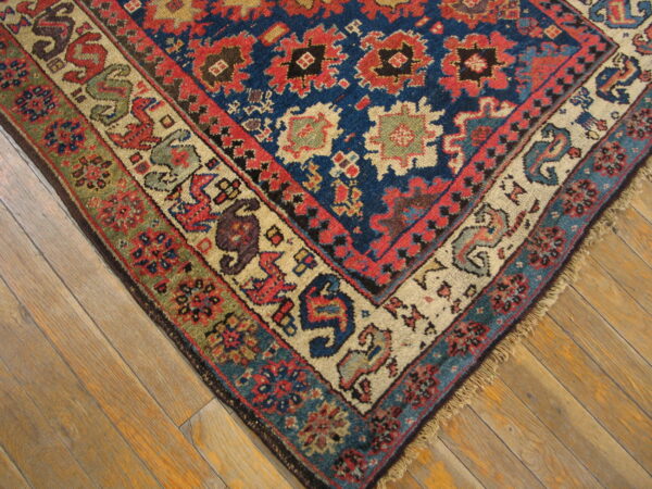 Low-pile tribal rug corner featuring blue, red, tan, and green patterns rests on wooden floorboards.