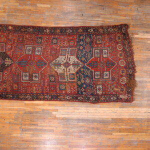 Distressed rectangular rug with complex geometric patterns in red, navy blue, and beige on wood flooring.