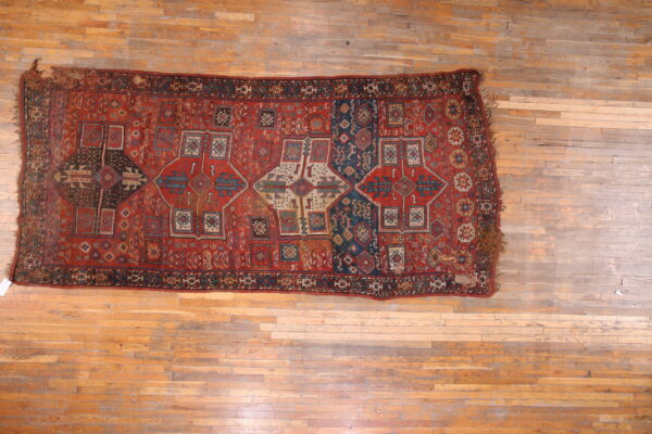 Distressed rectangular rug with complex geometric patterns in red, navy blue, and beige on wood flooring.