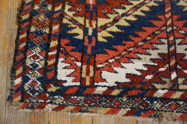 A shaggy rug corner with a stepped geometric pattern in red, blue, and white resting on wood flooring.