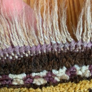 Detail view of a textured rug edge with long cream fringe and shag pile in purple, brown, and yellow.