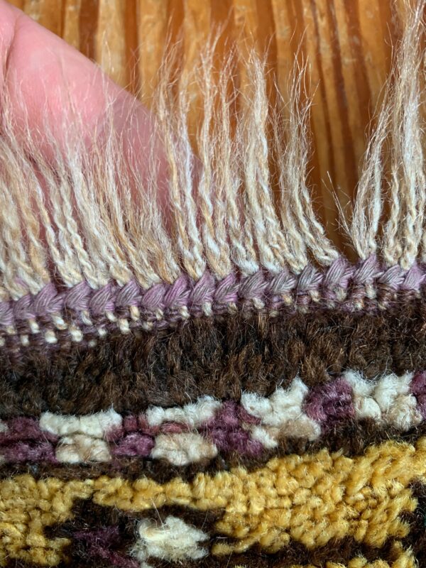 Detail view of a textured rug edge with long cream fringe and shag pile in purple, brown, and yellow.