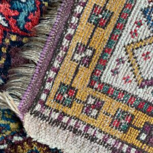 Detail of a yellow, cream, and purple geometric rug border overlapping a dense, dark red and blue floral rug.