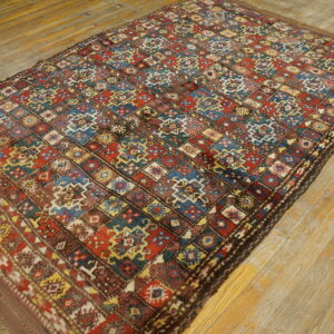 Low-pile rug with a complex, colorful diamond pattern in red, blue, and brown rests on distressed light hardwood.