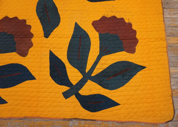 Mustard yellow quilted textile featuring dark teal and maroon floral motifs on weathered wood planks.