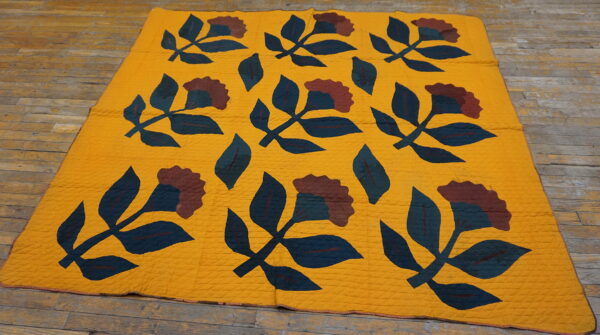 Flat textile in mustard yellow features repeating dark teal and brown floral quilt patterns on wood flooring.
