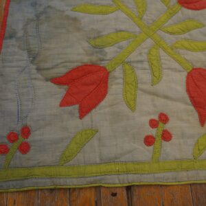 A flat, quilted textile featuring red and green floral appliqué lies on distressed hardwood flooring.