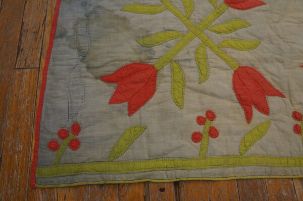 A flat, quilted textile featuring red and green floral appliqué lies on distressed hardwood flooring.