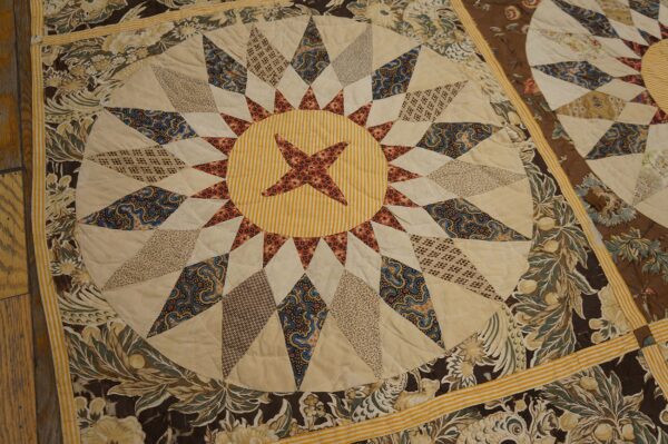 Close-up of a quilted textile with a central star medallion, surrounded by dark brown and cream floral borders on wood flooring.