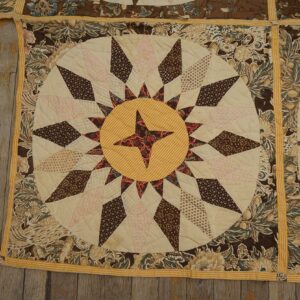 Quilted textile squares featuring brown floral borders and a central yellow and cream starburst pattern on wooden planks.