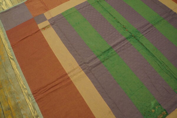 Quilted rug with thick stripes in rust, purple, green, and tan colors resting on distressed wood floors.