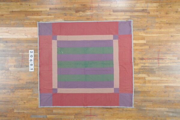 Block-patterned quilted textile in red, purple, and green, laid flat on worn wood plank flooring.