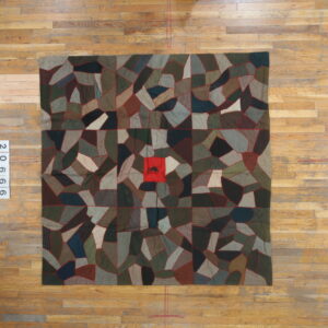 Muted brown and green patchwork rug with red seams and a central red square featuring a silhouette, laid on wood planks.