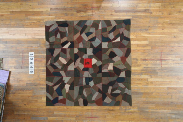 Muted brown and green patchwork rug with red seams and a central red square featuring a silhouette, laid on wood planks.