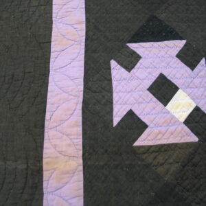Close up of dark quilted fabric with lavender and white geometric patchwork details and visible stitching.