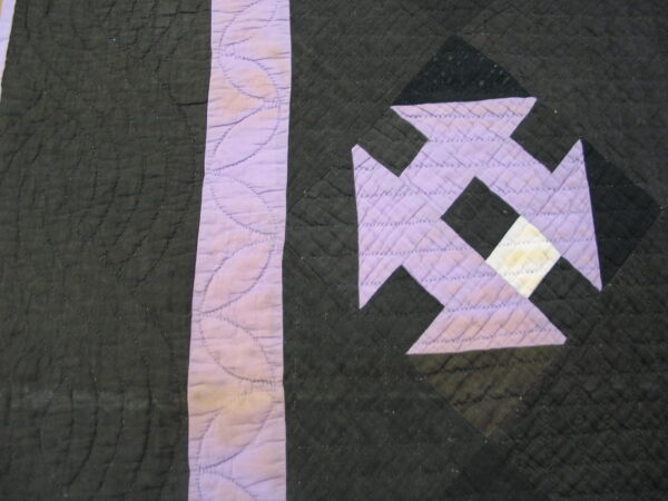 Close up of dark quilted fabric with lavender and white geometric patchwork details and visible stitching.
