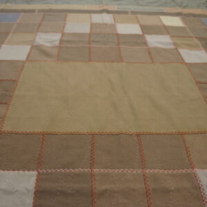 Low-pile patchwork textile in brown, beige, and grey squares joined by visible red and pink stitching.
