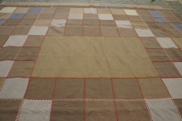 Low-pile patchwork textile in brown, beige, and grey squares joined by visible red and pink stitching.